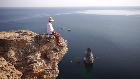 Digital Nomad Man in the Hat a Businessman with a Laptop Sits on the Rocks By the Sea During Sunset