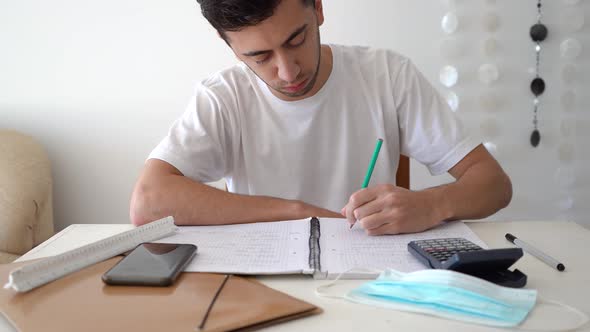 Male Student Writing Mathematical Formulas And Solutions On A Notebook With Calculator, Smartphone,