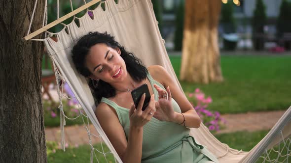 Portraitof a Woman Relaxing in a Hammock Outdoors Surfing Internet in Her Smartphone Clicks on the