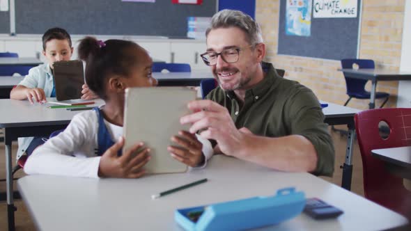 Diverse male teacher helping a schoolgirl sitting in classroom using tablet