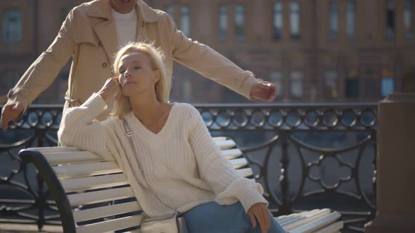 Senior Man Coming Behind and Closing Eyes of Woman Sitting on Bench By Riverside