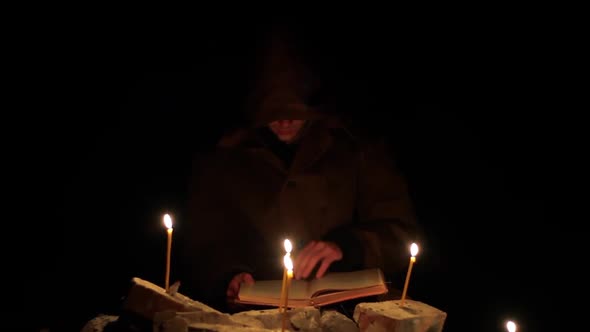 a Man in a Cloak with a Hood is Reading a Hot Book with Candles on the Floor