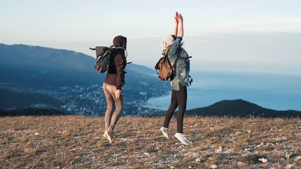 Two Young Happy Women Dancing and Raising Arms on Beautiful Mountain Top Over Sunset Sky