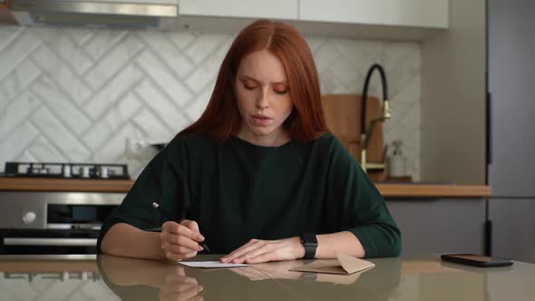 Serious Young Attractive Woman Writing Handwritten Letter and Puts It Away in Envelope Sitting at