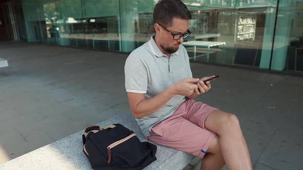 Middle Aged Man Is Swiping on Screen of Modern Mobile Phone, Sits on Street