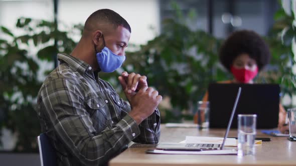 Diverse business people wearing face masks sitting using laptops going through paperwork in office