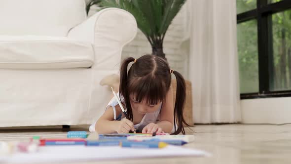 Cute little Asian girl lying on the floor drawing and painting on paper at home.