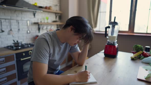 Side View of Concentrated Millennial Man Writing Ideas in Sketchpad with Pen Sitting at Kitchen