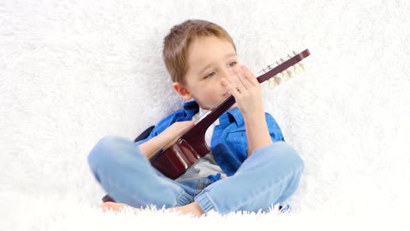 A Child Plays a Child's Acoustic Guitar with Pleasure While Sitting on a White Sofa