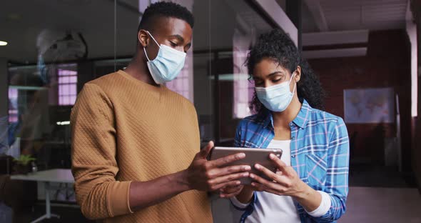 Diverse female and male business colleagues wearing masks in discussion looking at digital tablet