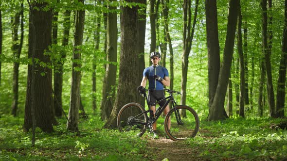 A Cyclist with a Smartphone in His Hands is Standing in the Middle of the Forest