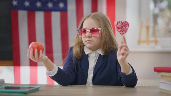 Thoughtful Teen Schoolgirl Choosing Apple or Lollipop Sitting in School Classroom at Desk