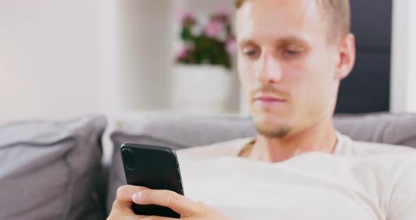 Happy Young Man Uses Smartphone While Sitting on a Sofa at Home Man Browses Through Internet Watches