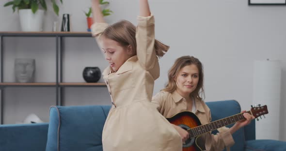 Charming Little Girl is Whirling and Dancing in Living Room Adult Woman is Playing Music on Guitar