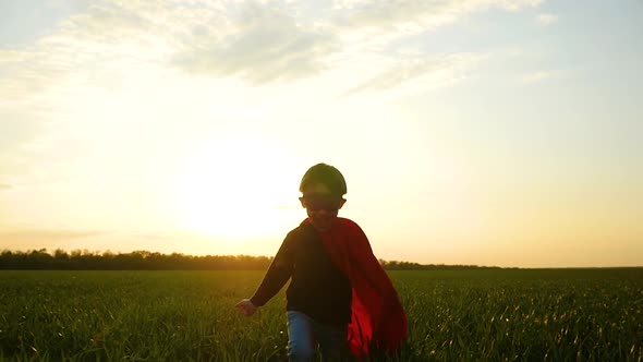 A Joyful Child in a Superhero Costume Runs Across the Green Grass To the Camera on a Sunset