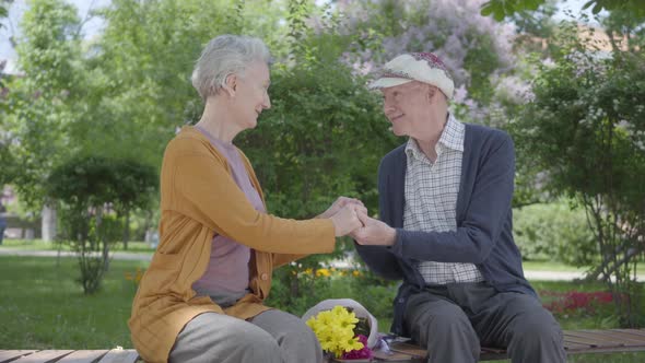 Old Woman with Bouquet of Yellow Flowers Sitting with an Old Man and Holding Hands in the Bench