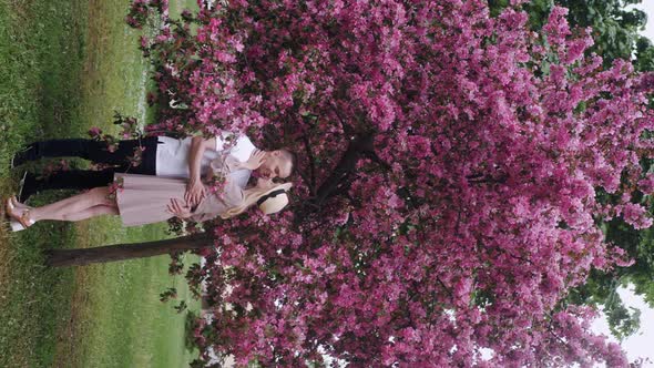 Romantic Beautiful Woman in a Hat Stands with a Man at a Cherry Blossom Tree in the Summer in the