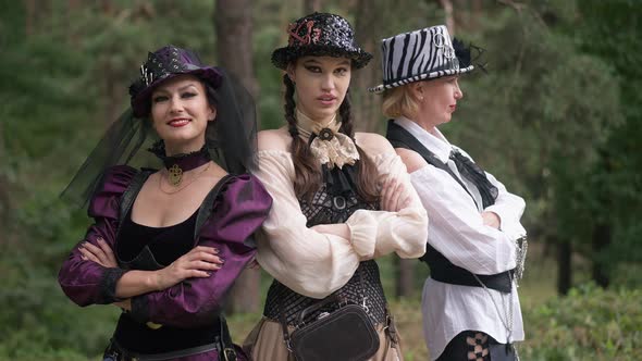 Three Confident Witches in Steampunk Costumes Crossing Hands Looking at Camera Standing in Forest on