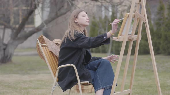 Pretty Young Smoking Girl Painting on Canvas While Sitting in the Backyard Outdoors