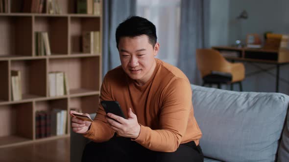 Happy Middle Aged Asian Man Making Online Money Transaction with Credit Card and Smartphone App Slow