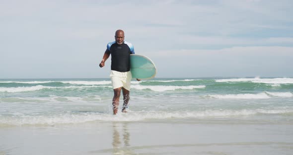 African american senior man walking on a beach holding surfboard and running out of the sea