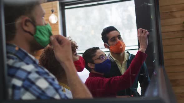 Diverse colleagues wearing face masks writing on glass board discussing together at modern office