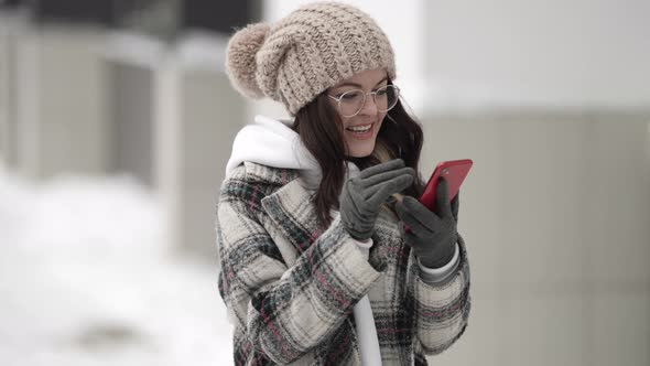 Smiling Teen Girl Is Using Mobile Phone on Street at Winter Swiping on Screen and Smiling