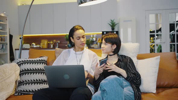 Attractive Lesbian Couple Sitting Side By Side on the Couch at Home Using Each of Their Gadgets