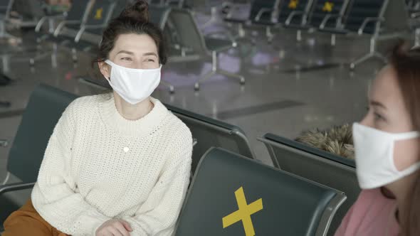 Two Women Tourists in Medical Mask Sitting in Airport Terminal at Safe Distance and Talking Before