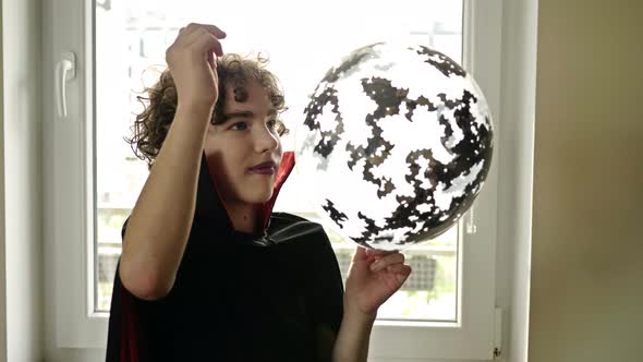 Halloween. Teenage Boy in a Vampire Costume Pierces a Balloon with Bats Confetti. Guy Is Delighted.