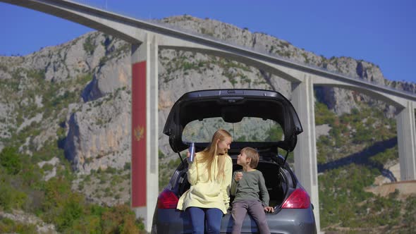 A Man and His Son are Sitting and Eating in Their Car with the Moracica Bridge in the Background