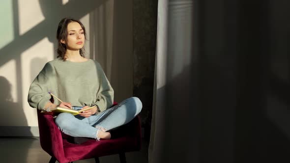 Beautiful Woman Sitting Near Window and Writing Things on Background of Sunset