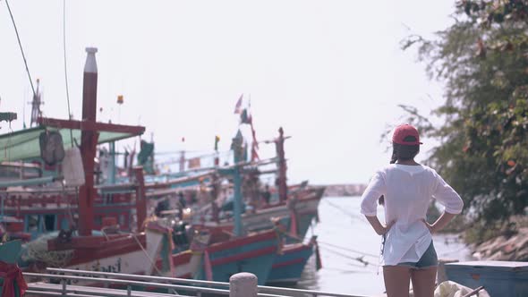 Girl in Long White Shirt and Red Cap Looks at Moored Boats