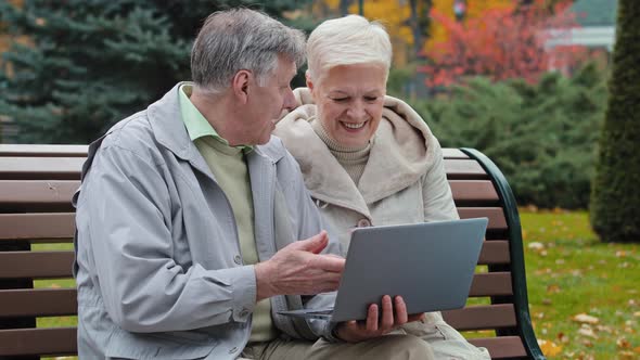 Happy Carefree Elderly Senior Grandparents Sit on Bench in Autumn Park Man Typing on Laptop Together