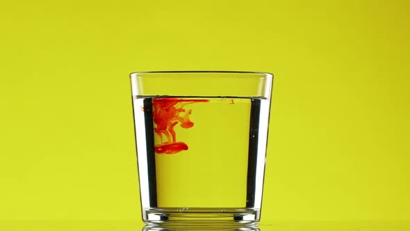 Drop of Red Ink Dripping Into Glass with Clear Water on Yellow Background in Slowmotion