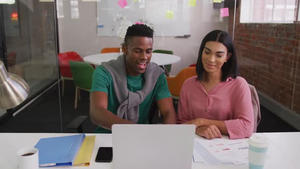 Diverse male and female business colleagues sitting at desk using laptop and smiling