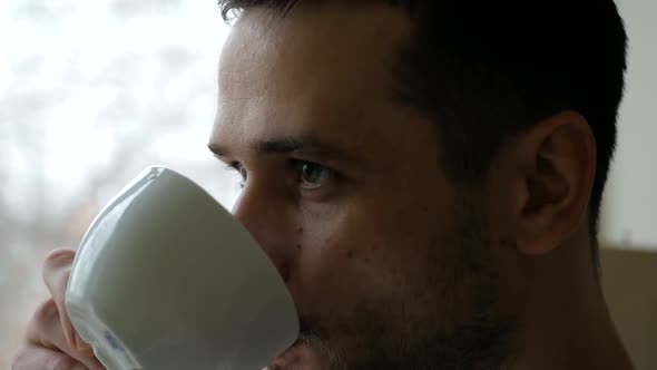 Closeup of a Young Man Standing in His Kitchen Drinking Coffee From a White Cup