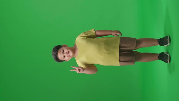 Full Body Of A Smiling Asian Little Boy Showing Gesture Peace While Standing On Green Screen alt