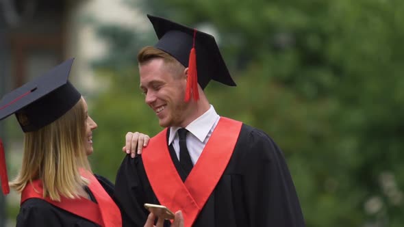 Couple of Graduates Laughing, Viewing Photos on Smartphone, Looking Into Future