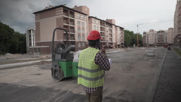 Builder in Protective Work Clothing Walks Through Construction of Houses Talking on Phone with