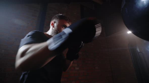 Side View of the Strong Boxer Man Hitting the Boxing Bag Wearing Black Boxing Gloves in the Boxing