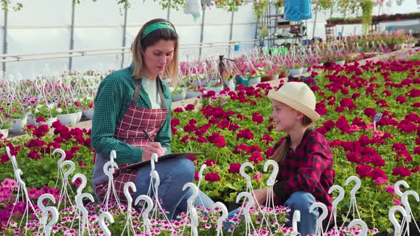 Kid with Woman Florist Working in Greenhouse with Notebook Examines the Growing Flowers