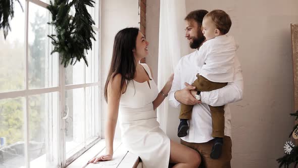Young Father is Holding His Baby Boy Standing By the Window While Attractive Mother in White Dress