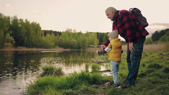 Curious Little Boy is Learning to Fish on Coast of Forest Lake Grandfather is Helping to Grandson