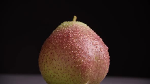 A large ripe yellow-red pear with water drops rotates around its axis on a dark background