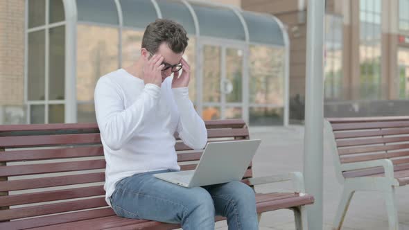 Young Man with Headache Using Laptop While Sitting Outdoor on Bench