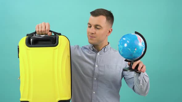 Happy Positive Man in a Blue Shirt Raises a Globe and a Yellow Suitcase to Shoulder Level and Smiles