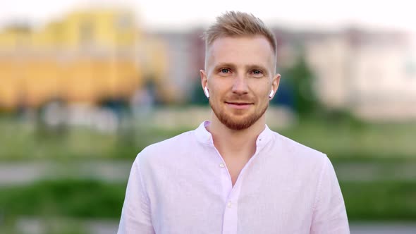 Adorable Young Smiling Male in Wireless Earphones Walking in Summer Park at City Background