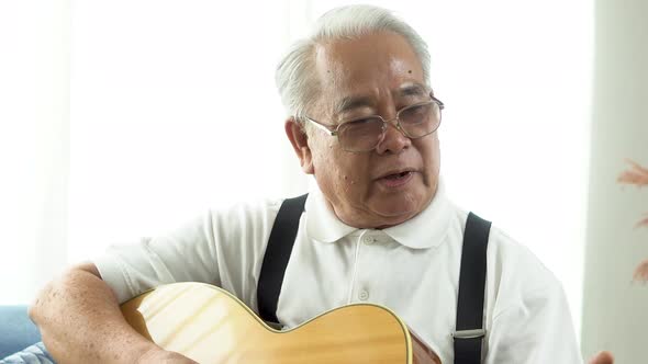 Asian Senior Man on Sofa and Playing Acoustic Guitar