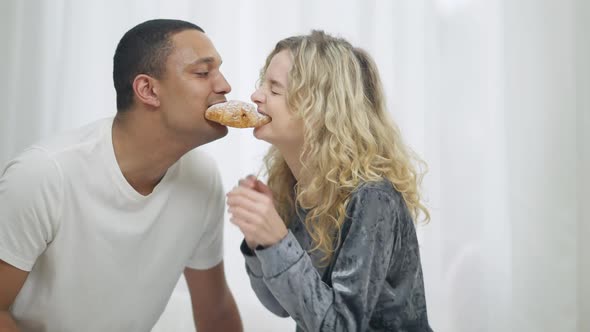 Side View of Cheerful Millennial Interracial Couple Eating Croissant From Both Sides in Bedroom in